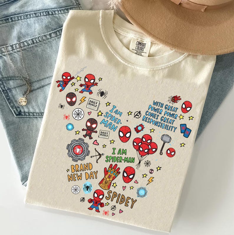 Puede incluir: Camiseta color crema con un colorido gr&aacute;fico de Spider-Man. El dise&ntilde;o incluye varias ilustraciones de Spider-Man, el texto "I am Spider-Man" y otros s&iacute;mbolos relacionados. La camiseta est&aacute; doblada sobre un par de jeans.