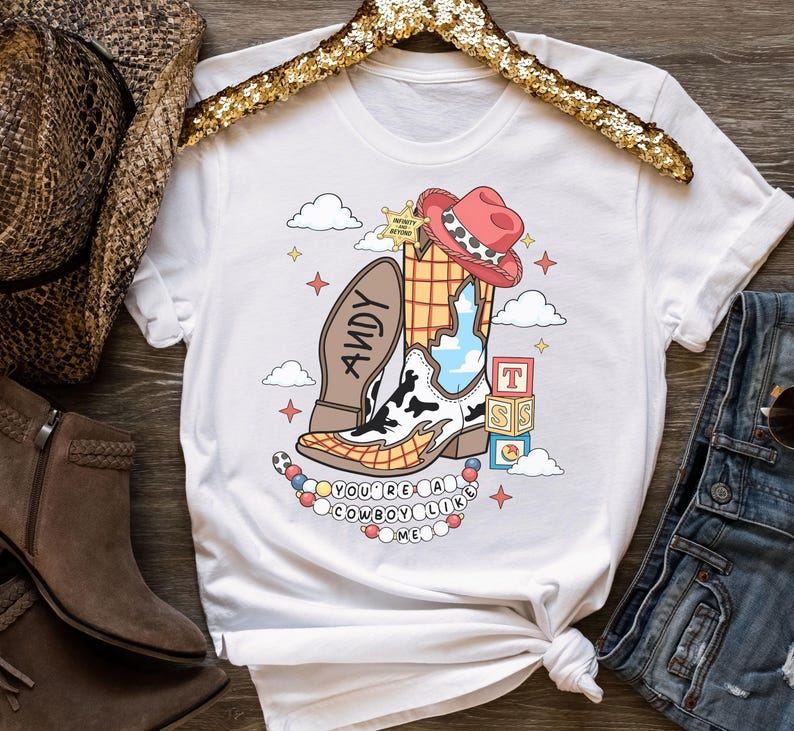 May include: A white t-shirt featuring a graphic print of a cowboy boot with a cow-print pattern, a red hat, a sheriff badge with the text Infinity and Beyond, alphabet blocks, and the text Andy on the sole. The text reads You're a cowboy like me.