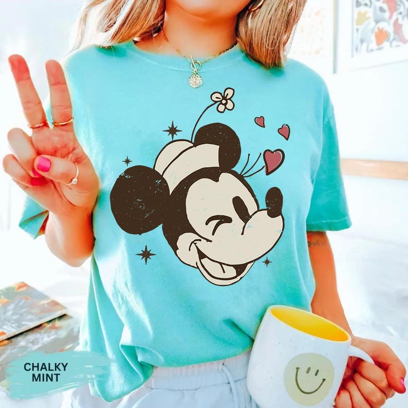 May include: A light blue t-shirt featuring a smiling Mickey Mouse graphic with hearts and stars. The shirt has the words "CHALKY MINT" printed on it. A white mug with a yellow interior and a smiley face is held in the hand.