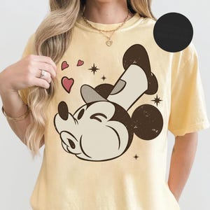 May include: A light yellow t-shirt featuring a distressed graphic of Mickey Mouse with hearts and stars. The cartoon character is in a relaxed pose, with a hat and a smiling expression. The shirt has a classic, vintage style.