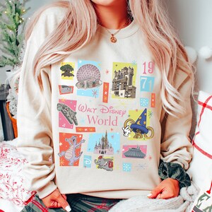 May include: Beige sweatshirt featuring colorful squares with Disney World icons and the text "Walt Disney World" and "1971". The design includes images of the Millennium Falcon, a dragon, and Cinderella's castle.