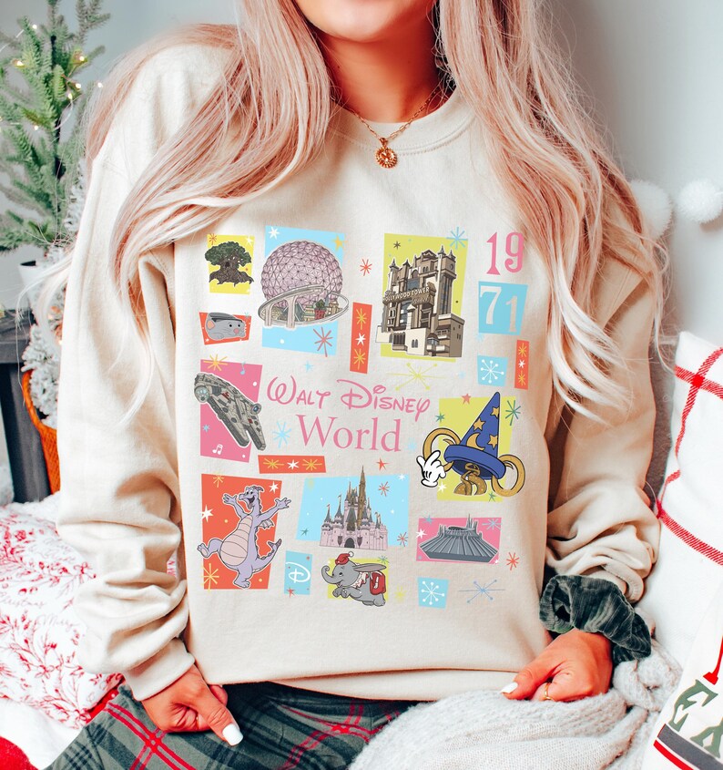May include: Beige sweatshirt featuring colorful squares with Disney World icons and the text "Walt Disney World" and "1971". The design includes images of the Millennium Falcon, a dragon, and Cinderella's castle.
