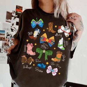 May include: Dark gray t-shirt featuring colorful cartoon illustrations of Toy Story characters' accessories, including boots, bows, and a green bow. The names of the characters are written below each illustration. The shirt has a crew neck.