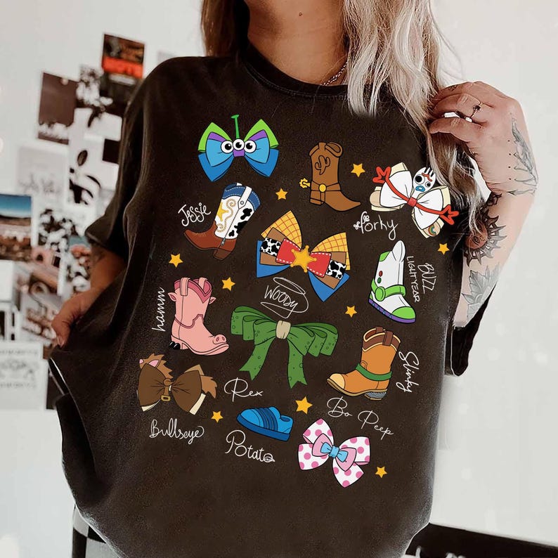 May include: Dark gray t-shirt featuring colorful cartoon illustrations of Toy Story characters' accessories, including boots, bows, and a green bow. The names of the characters are written below each illustration. The shirt has a crew neck.
