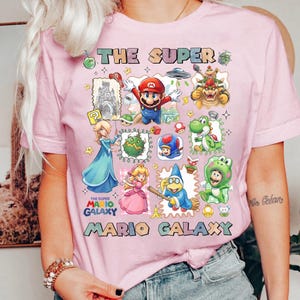 May include: Pink t-shirt featuring a colorful graphic of characters and elements from the Super Mario Galaxy video game. The design includes Mario, Princess Peach, Bowser, and other iconic figures, with the text "THE SUPER MARIO GALAXY".