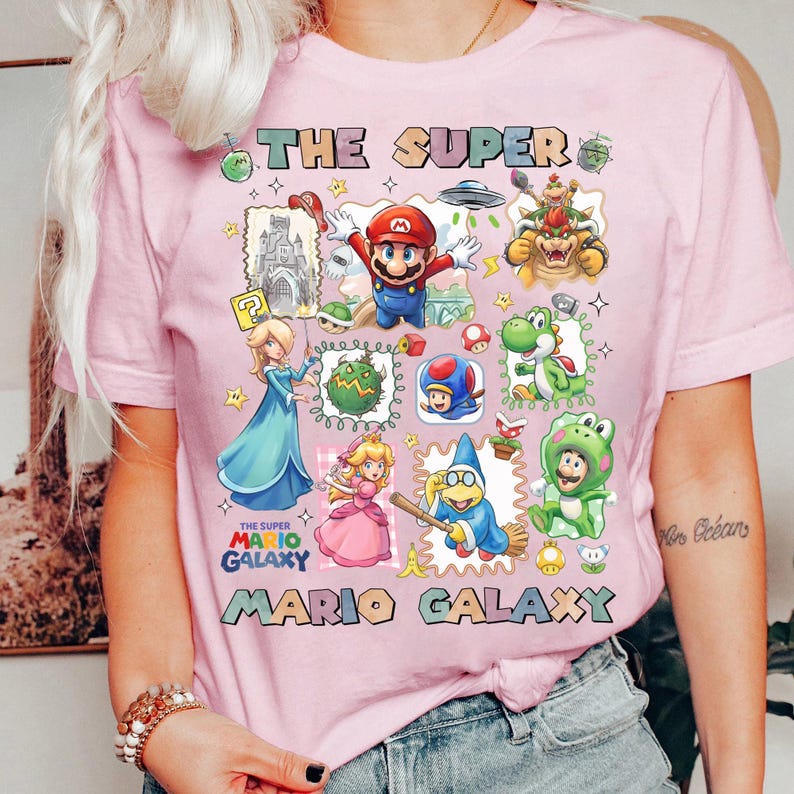 May include: Pink t-shirt featuring a colorful graphic of characters and elements from the Super Mario Galaxy video game. The design includes Mario, Princess Peach, Bowser, and other iconic figures, with the text "THE SUPER MARIO GALAXY".