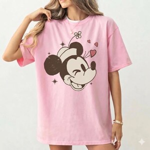 May include: Pink t-shirt featuring a smiling Mickey Mouse graphic with hearts and stars. The graphic is in brown and cream colors. The shirt has short sleeves and a crew neck.