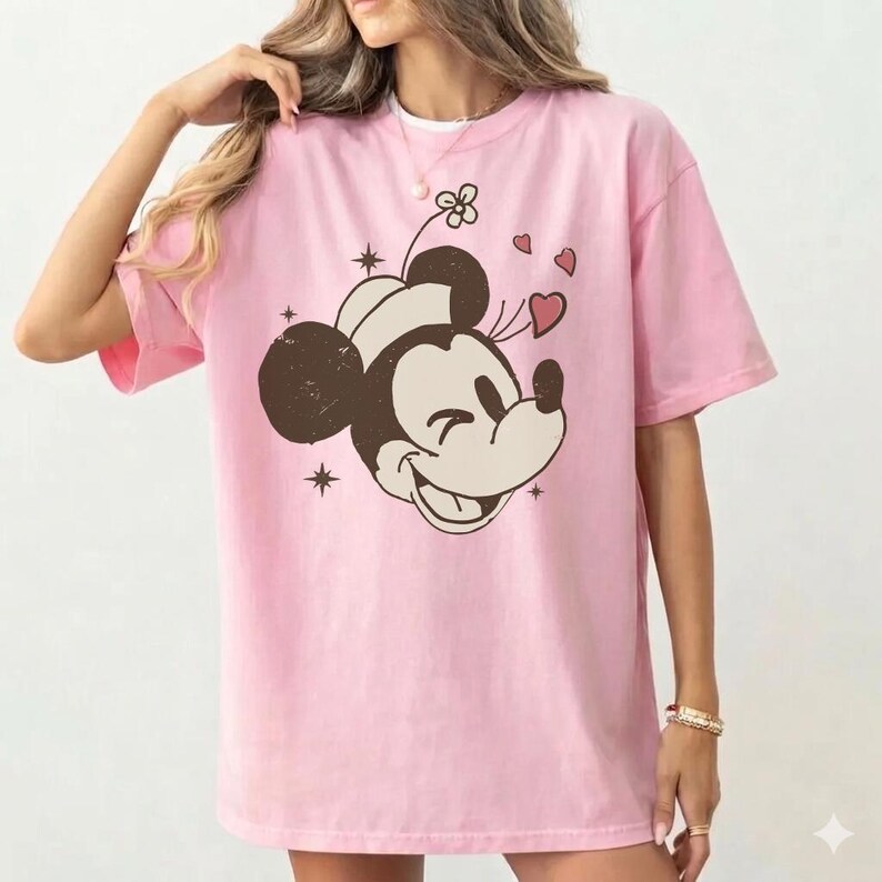May include: Pink t-shirt featuring a smiling Mickey Mouse graphic with hearts and stars. The graphic is in brown and cream colors. The shirt has short sleeves and a crew neck.