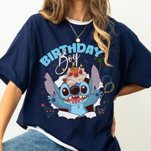 K&ouml;nnte beinhalten: Marineblaues T-Shirt mit einem Cartoon-Charakter und dem Text "BIRTHDAY Boy". Die Figur hat einen Geburtstagskuchen auf dem Kopf. Das Design umfasst Feuerwerk, Luftballons und eine Partyh&uuml;tchen.