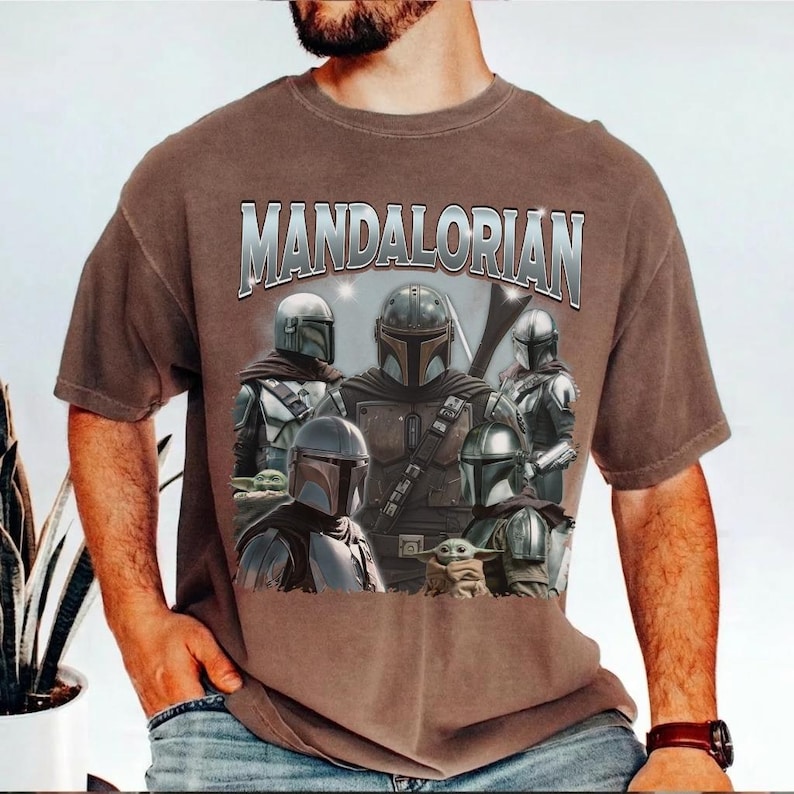May include: Brown t-shirt featuring a graphic of The Mandalorian characters and the text "MANDALORIAN" in silver. The design includes multiple figures in armor and a small green creature. The shirt is a short-sleeved style.