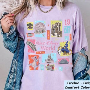 May include: Orchid-colored t-shirt with a colorful graphic design featuring various Walt Disney World icons, including the Millennium Falcon, Cinderella's Castle, and the Sorcerer's Hat. The text "Walt Disney World" and the year "1971" are also displayed.