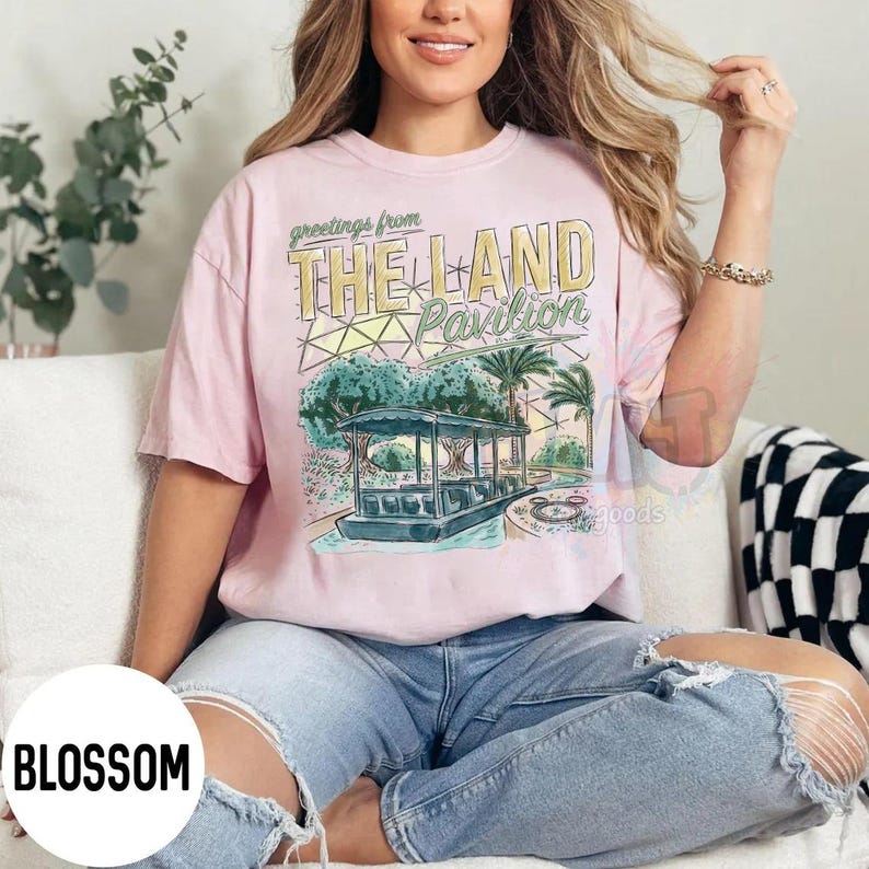 Puede incluir: Una camiseta de manga corta de color rosa claro con un estampado gr&aacute;fico que incluye el texto Greetings from THE LAND Pavilion. El dise&ntilde;o muestra una ilustraci&oacute;n estilizada de un barco en el agua, palmeras y una c&uacute;pula geom&eacute;trica en tonos suaves.