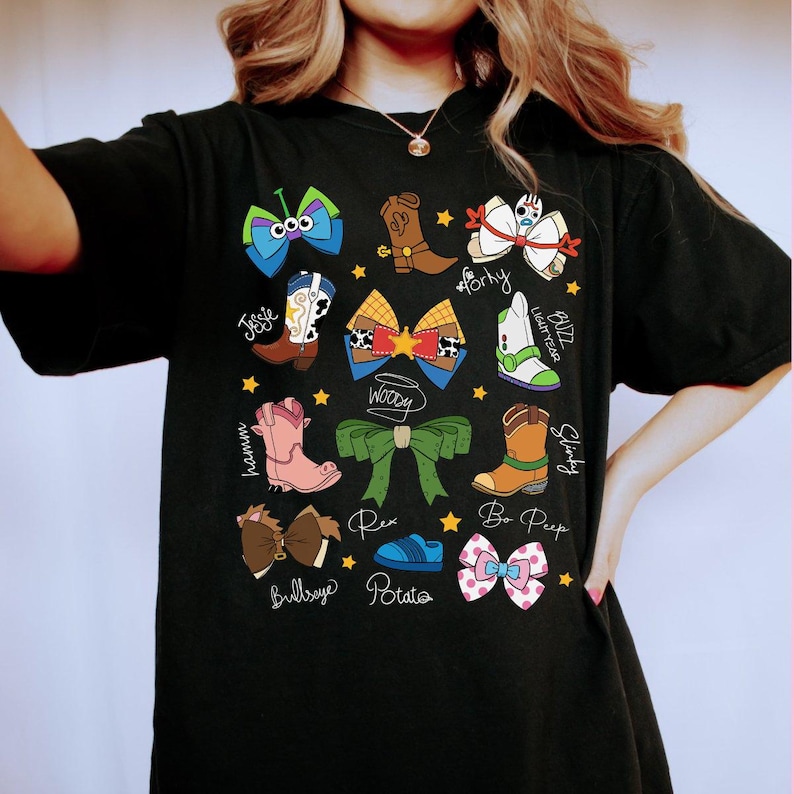 May include: Black t-shirt featuring colorful cartoon bow designs with character names like Woody, Jessie, and Buzz Lightyear. The bows are in various colors and styles, with star accents.