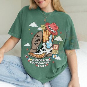 May include: A green short-sleeve t-shirt featuring a graphic of a cowboy boot with a cow print pattern, a red hat, a sheriff star, and alphabet blocks. The text reads "ANDY" on the boot sole and "YOU'RE A COWBOY LIKE ME" below the boot.