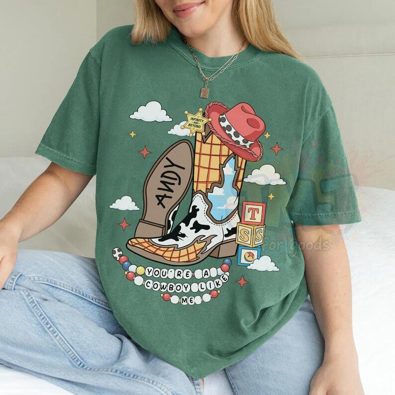 May include: A green short-sleeve t-shirt featuring a graphic of a cowboy boot with a cow print pattern, a red hat, a sheriff star, and alphabet blocks. The text reads "ANDY" on the boot sole and "YOU'RE A COWBOY LIKE ME" below the boot.