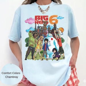 May include: Light blue t-shirt featuring a colorful graphic of characters and the text "Big Hero 6". The design includes a cityscape, clouds, and the number 6. The shirt is a Comfort Colors Chambray.