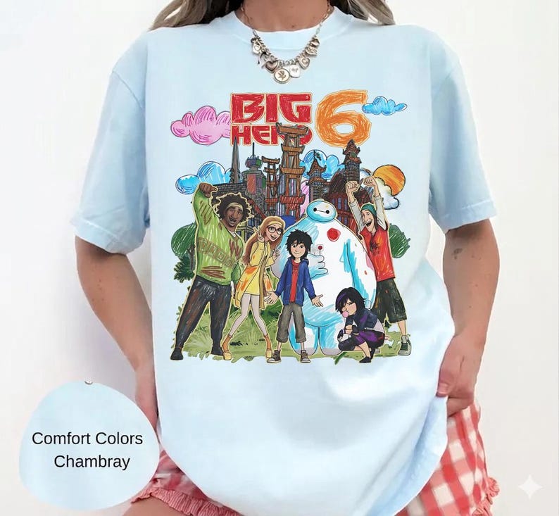 May include: Light blue t-shirt featuring a colorful graphic of characters and the text "Big Hero 6". The design includes a cityscape, clouds, and the number 6. The shirt is a Comfort Colors Chambray.
