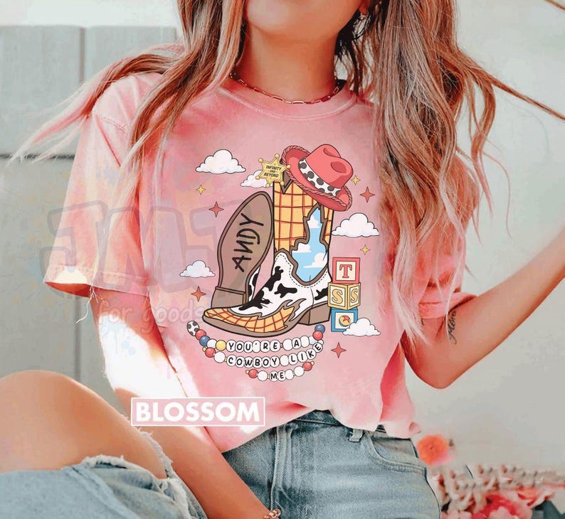 May include: A pink short-sleeve t-shirt featuring a graphic of a cowboy boot with a cow print and yellow grid pattern, a red cowboy hat, a sheriff badge, alphabet blocks, and the text "ANDY" and "YOU'RE A COWBOY LIKE ME".