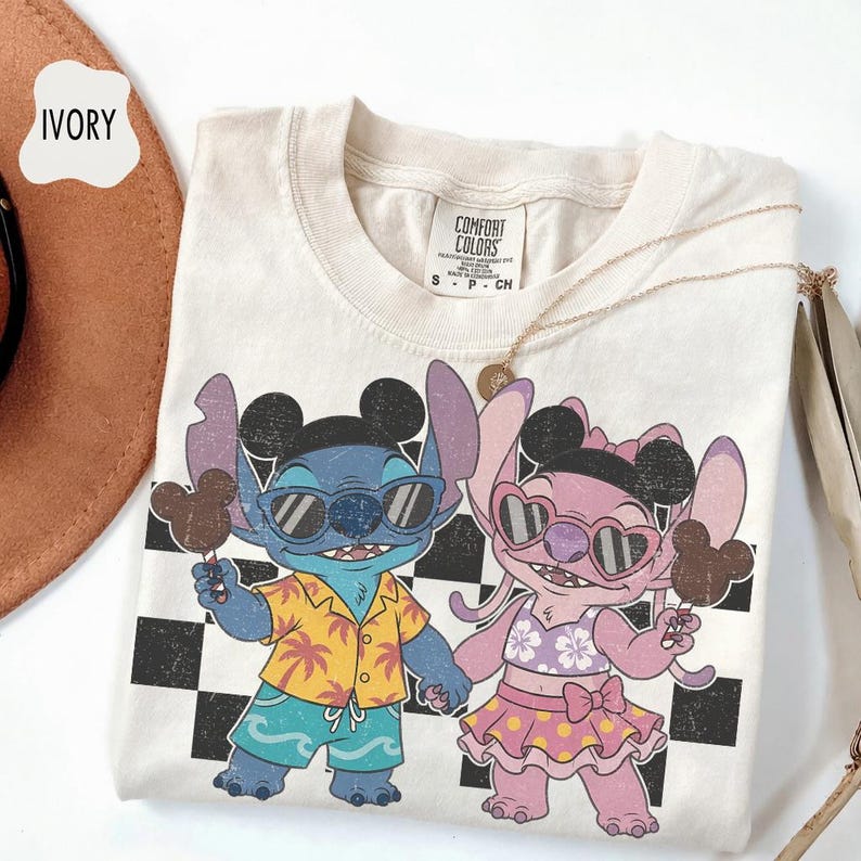 May include: Ivory-colored t-shirt featuring a cartoon illustration of Stitch and Angel holding Mickey Mouse-shaped ice cream. Stitch wears sunglasses, a yellow shirt, and blue shorts. Angel wears sunglasses and a pink skirt. The shirt has a checkered background.