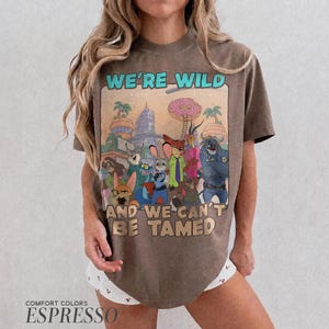 K&ouml;nnte beinhalten: Ein braunes kurz&auml;rmeliges T-Shirt mit einem Grafikdruck verschiedener animierter Tierfiguren aus Zootopia. Der Text &uuml;ber den Figuren lautet WE'RE WILD und der Text darunter lautet AND WE CAN'T BE TAMED. Das Shirt hat einen Rundhalsausschnitt.