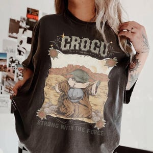 May include: Dark gray t-shirt featuring a graphic of a character with the text "GROGU" above the image and "STRONG WITH THE FORCE" below. The graphic is set against a desert landscape with a brown and tan color palette.