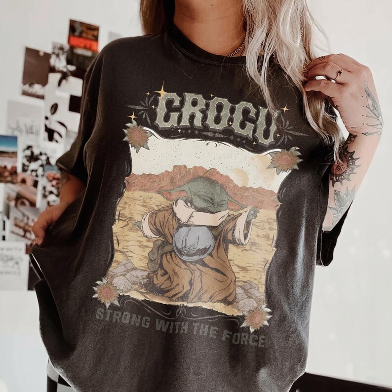 May include: Dark gray t-shirt featuring a graphic of a character with the text "GROGU" above the image and "STRONG WITH THE FORCE" below. The graphic is set against a desert landscape with a brown and tan color palette.