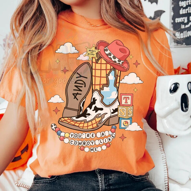 May include: An orange t-shirt featuring a graphic print of a cowboy boot with a cow print pattern, a red hat, a sheriff badge labeled Infinity and Beyond, alphabet blocks, and the text Andy on the sole and You've Got a Friend in Me lyrics.