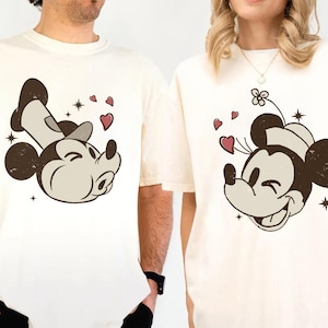 May include: Cream-colored t-shirts featuring a smiling Mickey Mouse graphic with hearts and stars. The Mickey Mouse design is in brown, beige, and white. The shirts have short sleeves and a relaxed fit.