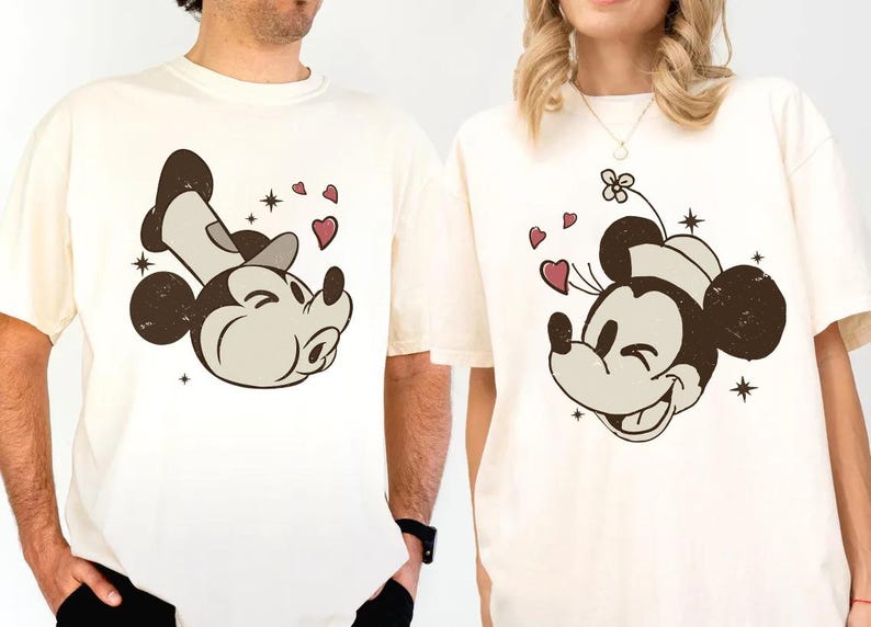 May include: Cream-colored t-shirts featuring a smiling Mickey Mouse graphic with hearts and stars. The Mickey Mouse design is in brown, beige, and white. The shirts have short sleeves and a relaxed fit.