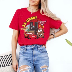 May include: Red t-shirt featuring a Cars-themed graphic with the text "KA-CHOW!" and the number 4. The design includes illustrations of Lightning McQueen and Mater. The shirt is worn by a person.