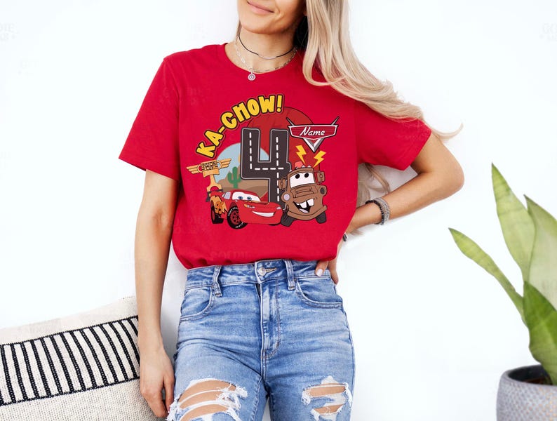 May include: Red t-shirt featuring a Cars-themed graphic with the text "KA-CHOW!" and the number 4. The design includes illustrations of Lightning McQueen and Mater. The shirt is worn by a person.