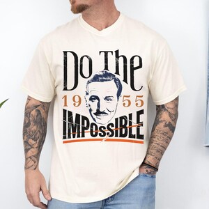 Pu&ograve; includere: T-shirt color crema con un design grafico. Il design presenta le parole "Do The Impossible" in nero, con gli anni "1955" in arancione. Un ritratto di un uomo &egrave; al centro. La maglietta ha uno scollo a V.