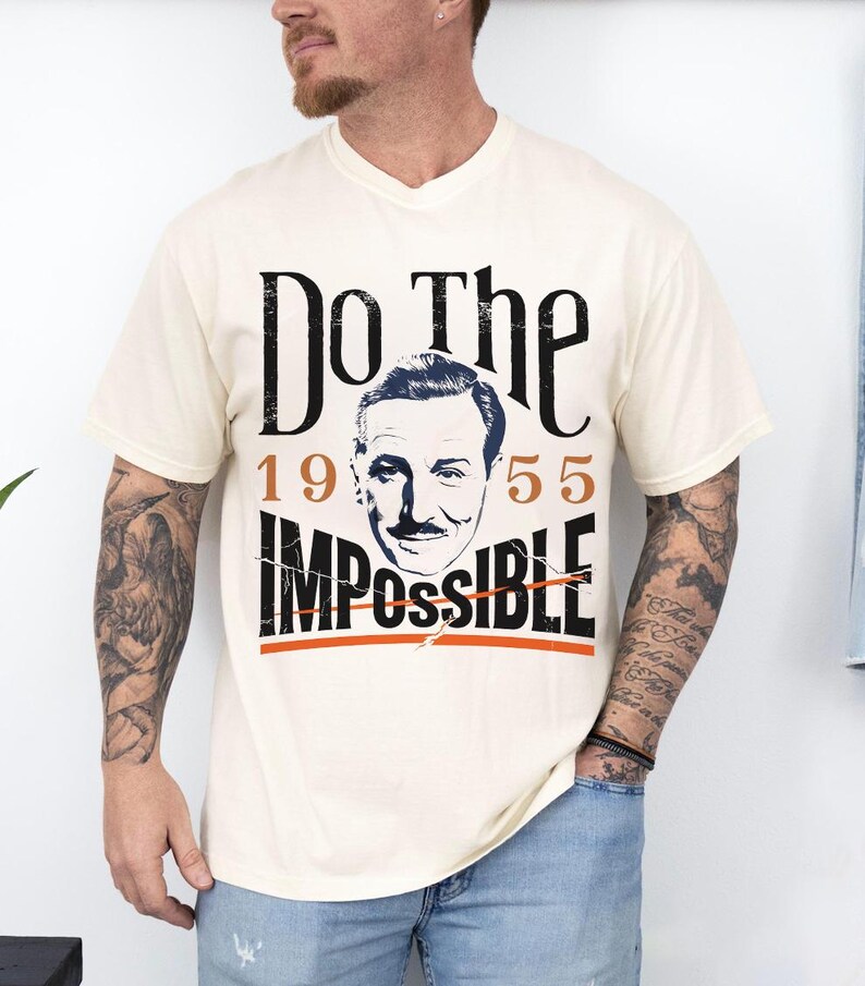 Pu&ograve; includere: T-shirt color crema con un design grafico. Il design presenta le parole "Do The Impossible" in nero, con gli anni "1955" in arancione. Un ritratto di un uomo &egrave; al centro. La maglietta ha uno scollo a V.