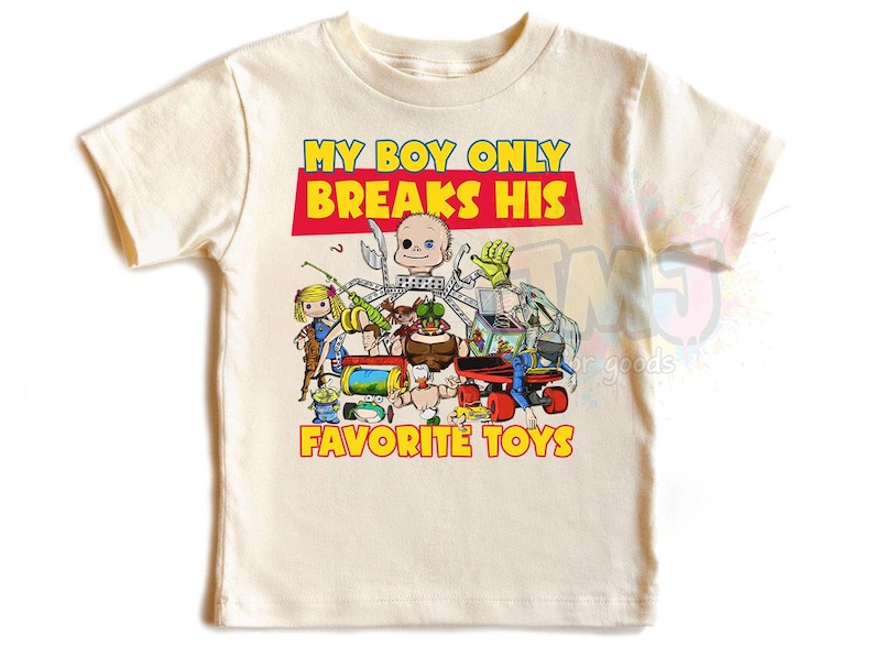 May include: A cream-colored short-sleeve t-shirt featuring a graphic print of various toys and a doll head. The text reads MY BOY ONLY BREAKS HIS FAVORITE TOYS in yellow and red lettering. The design includes a jack-in-the-box, a green alien, and a red car.