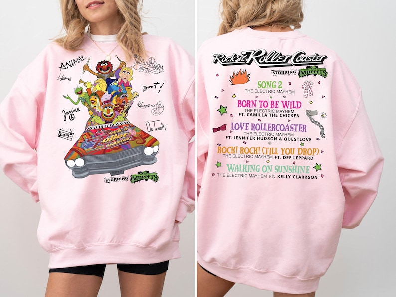 May include: Pink sweatshirt with a Muppets graphic on the front, including characters in a car. The back displays "Rockin' Roller Coaster" and song titles. The long-sleeved sweatshirt is a casual garment.