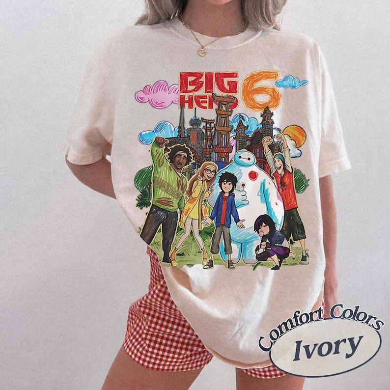 May include: Ivory-colored t-shirt featuring a colorful graphic of characters from "Big Hero 6." The design includes the text "BIG HERO 6" and a depiction of the characters in front of a cityscape. The shirt is a Comfort Colors brand.