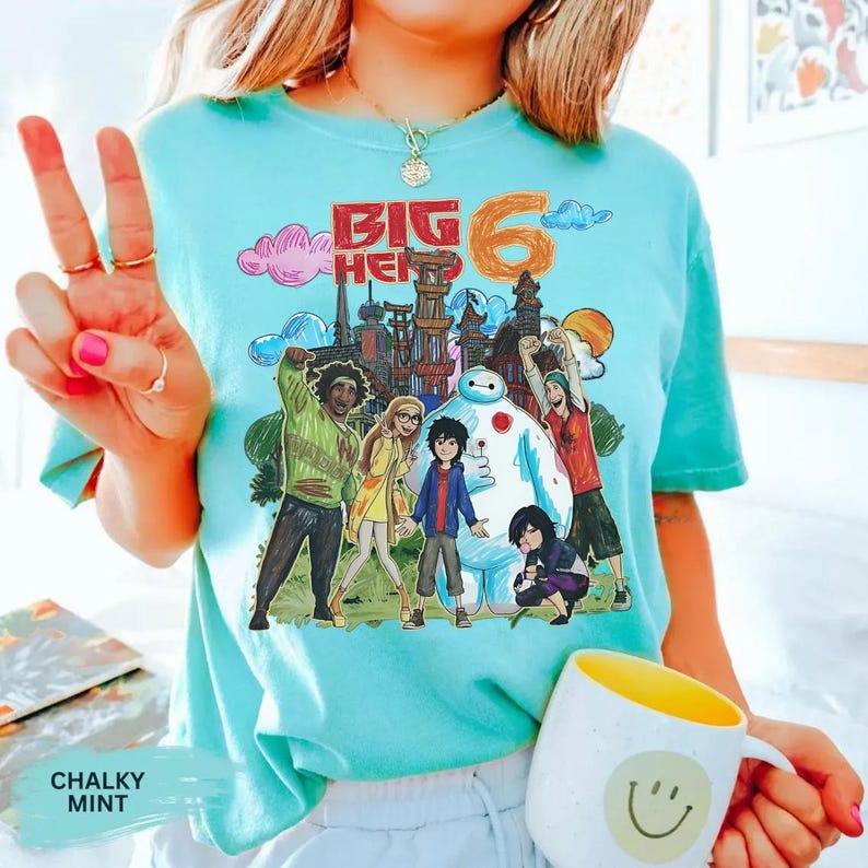 May include: A light blue t-shirt featuring a colorful graphic of the Big Hero 6 characters and the text "BIG 6". The design includes Baymax and other characters in front of a cityscape. A white mug with a yellow interior and a smiley face is also visible.