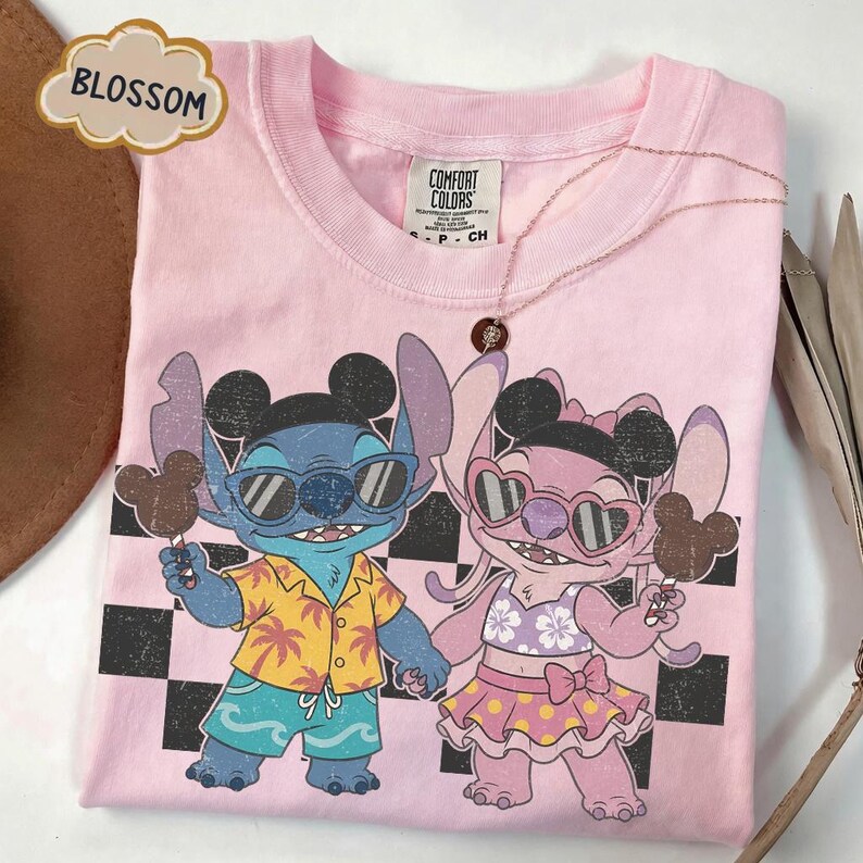 May include: A light pink t-shirt featuring a graphic of two cartoon characters holding ice cream. The characters are wearing sunglasses and tropical attire. The shirt has a black and white checkered pattern in the background and the word "BLOSSOM" in a cloud-shaped text.