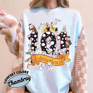 May include: Light blue sweatshirt featuring a graphic of the number 101 with Dalmatian dogs and characters from the Disney movie. The word "Dalmatians" is on a yellow banner. The sweatshirt has the words "Comfort Colors Chambray" on the bottom left.