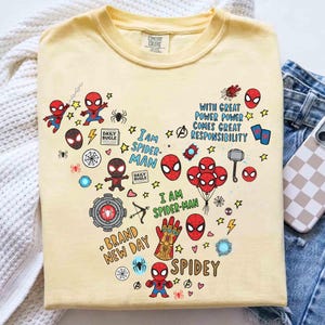 Puede incluir: Una camiseta amarillo claro con un dise&ntilde;o en forma de coraz&oacute;n con varios gr&aacute;ficos de Spider-Man. El dise&ntilde;o incluye figuras de Spider-Man, m&aacute;scaras y texto como "I AM SPIDER-MAN", "BRAND NEW DAY" y "SPIDEY". La camiseta se muestra con unos vaqueros azules y una funda de tel&eacute;fono.