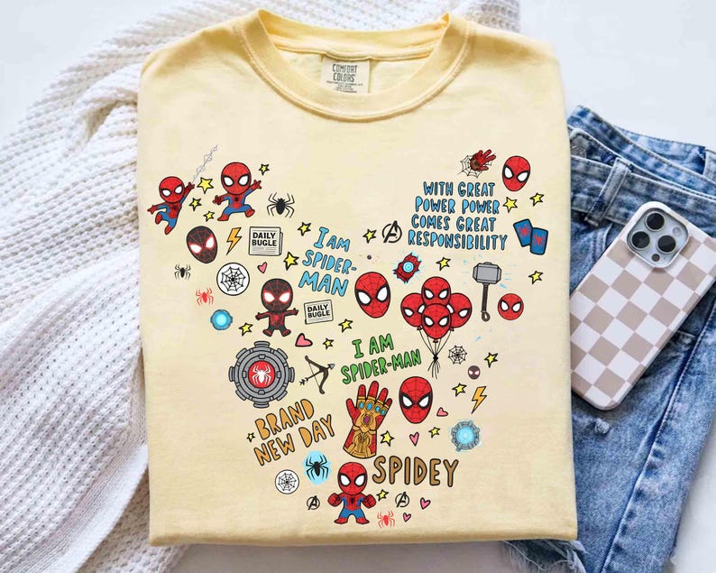 Puede incluir: Una camiseta amarillo claro con un dise&ntilde;o en forma de coraz&oacute;n con varios gr&aacute;ficos de Spider-Man. El dise&ntilde;o incluye figuras de Spider-Man, m&aacute;scaras y texto como "I AM SPIDER-MAN", "BRAND NEW DAY" y "SPIDEY". La camiseta se muestra con unos vaqueros azules y una funda de tel&eacute;fono.