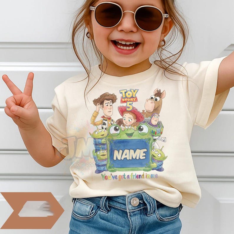 May include: A cream-coloured short-sleeve t-shirt featuring a colourful graphic print of Toy Story 5 characters including Woody, Jessie, Buzz Lightyear, and Bullseye surrounding a green frog holding a sign with the customisable text NAME.