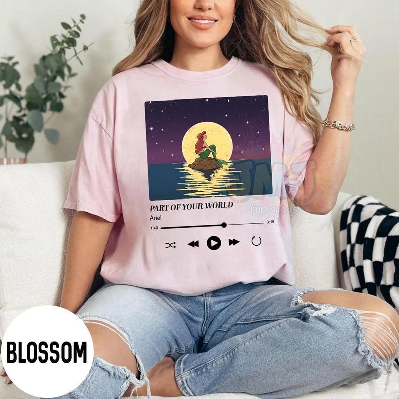 May include: A light pink short-sleeve t-shirt featuring a graphic of a mermaid sitting on a rock under a full moon. The design includes a music player interface with the text Part of Your World, Ariel, and playback controls for the song.