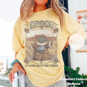 May include: A light yellow t-shirt with a graphic of Grogu from The Mandalorian. The graphic features Grogu in a brown robe, holding a silver ball, with the text "GROGU" and "STRONG WITH THE FORCE". The shirt is labeled "Comfort Colors Banana".