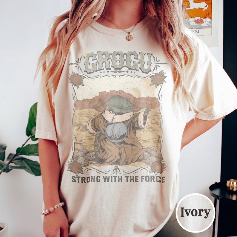 May include: Ivory-colored t-shirt featuring a Grogu graphic. The design includes the word "GROGU" above an illustration of Grogu, with the text "STRONG WITH THE FORCE" below. The shirt is a relaxed fit.