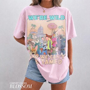 K&ouml;nnte beinhalten: Ein hellrosa T-Shirt mit kurzen &Auml;rmeln und einem Grafikdruck von animierten Zootopia-Figuren. Der Text lautet WE'RE WILD AND WE CAN'T BE TAMED. Das Design zeigt eine Stadtkulisse, einen gro&szlig;en Donut und verschiedene tierische Charaktere.