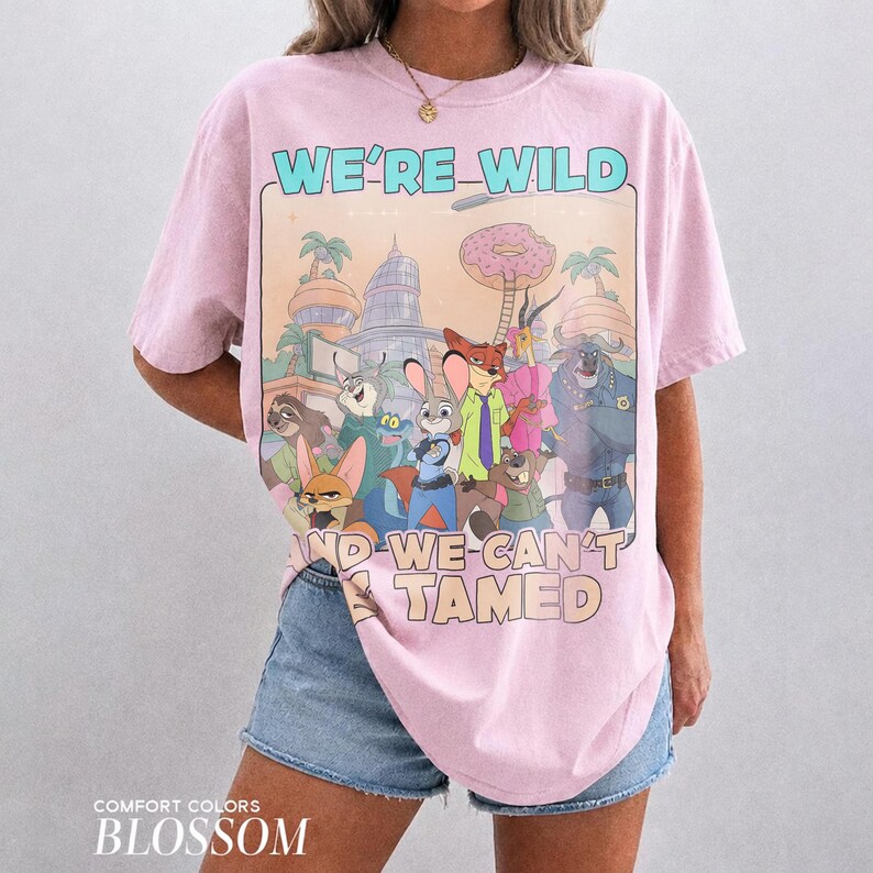 K&ouml;nnte beinhalten: Ein hellrosa T-Shirt mit kurzen &Auml;rmeln und einem Grafikdruck von animierten Zootopia-Figuren. Der Text lautet WE'RE WILD AND WE CAN'T BE TAMED. Das Design zeigt eine Stadtkulisse, einen gro&szlig;en Donut und verschiedene tierische Charaktere.