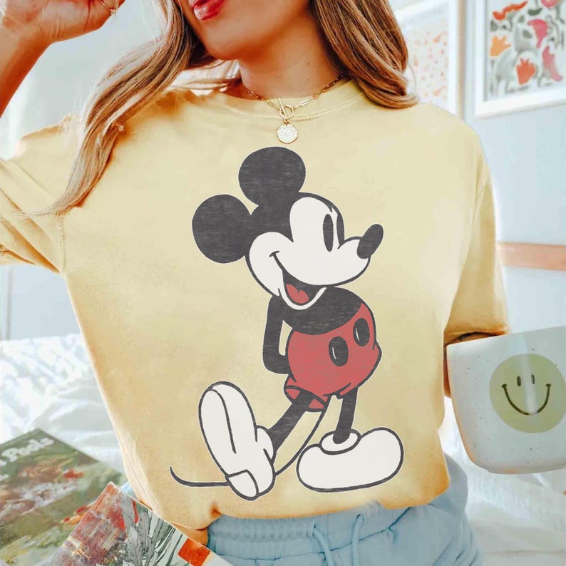 Peut inclure: Un t-shirt jaune clair avec un grand dessin de Mickey Mouse. Mickey est repr&eacute;sent&eacute; dans sa pose classique, avec des oreilles noires et un visage souriant. Son short est rouge et ses chaussures sont blanches. Le t-shirt est de style d&eacute;contract&eacute;.