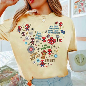 Puede incluir: Una camiseta amarilla clara con un dise&ntilde;o en forma de coraz&oacute;n con varios gr&aacute;ficos y textos de Spider-Man. El dise&ntilde;o incluye figuras de Spider-Man, m&aacute;scaras y frases como "I am Spider-Man" y "Brand New Day".