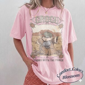 May include: Pink Comfort Colors t-shirt featuring a Grogu graphic with the text "STRONG WITH THE FORCE." The design includes a desert landscape and vintage-style lettering. The shirt is a light pink color.